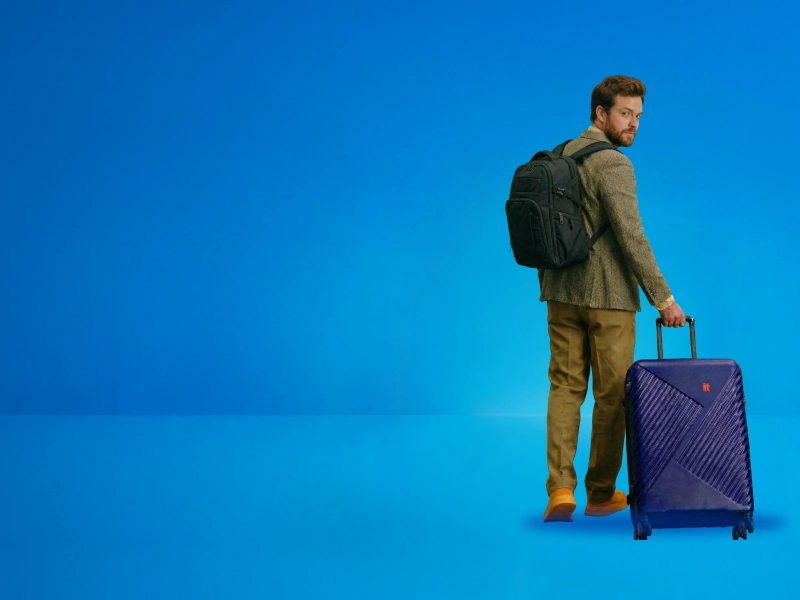 A man carrying a suitcase and a backpack looking backwards as he walks away